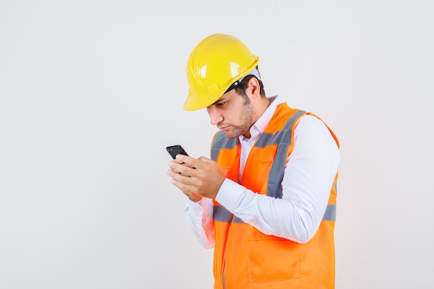 Homme de constructeur à l'aide de smartphone en chemise, uniforme et à la recherche de occupé. vue de face.