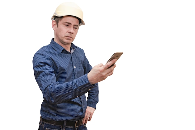 Un homme constructeur ou architecte dans un casque sur fond blanc isolé regarde l'écran d'un téléphone mobile.