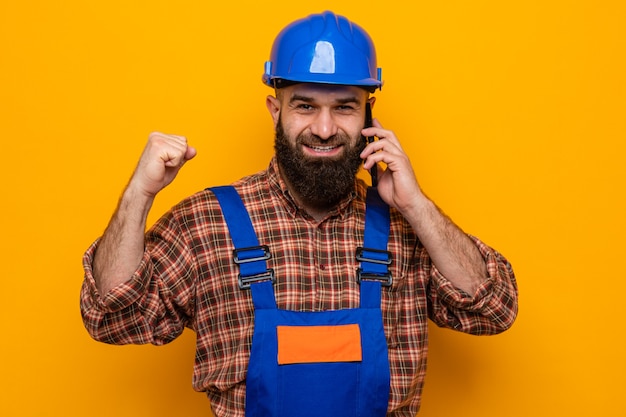 Homme constructeur barbu en uniforme de construction et casque de sécurité heureux et excité serrant le poing souriant tout en parlant au téléphone portable debout sur fond orange