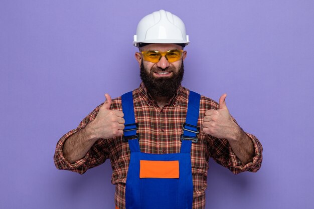 Photo gratuite homme constructeur barbu en uniforme de construction et casque de sécurité portant des lunettes de sécurité jaunes regardant la caméra souriant joyeusement montrant les pouces vers le haut debout sur fond violet