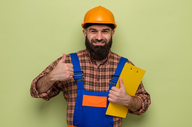 Homme constructeur barbu en uniforme de construction et casque de sécurité tenant le presse-papiers regardant la caméra souriant joyeusement montrant les pouces vers le haut debout sur fond vert