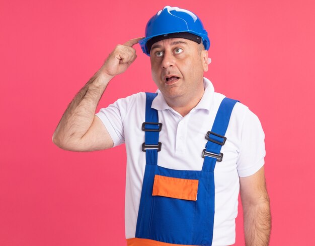 Homme de constructeur caucasien adulte impressionné en uniforme met le doigt sur un casque de sécurité sur rose