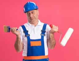 Photo gratuite homme de constructeur caucasien adulte insatisfait en uniforme tient un pinceau et un pinceau à rouleau sur rose
