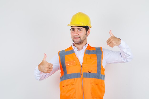 Homme de constructeur en chemise, uniforme montrant les pouces vers le haut et à la vue de face, heureux.