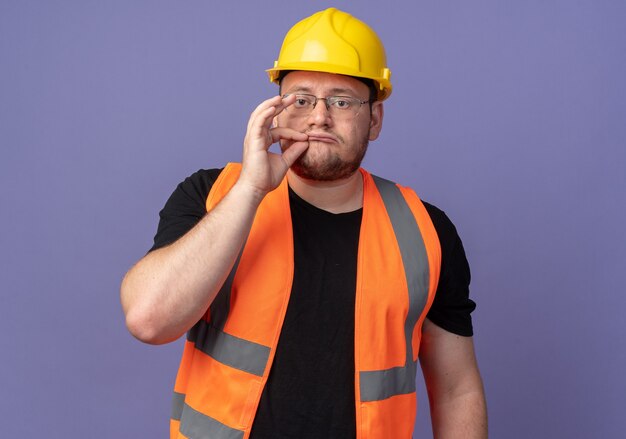Homme de constructeur en gilet de construction et casque de sécurité faisant un geste de silence avec les doigts comme fermer la bouche avec une fermeture à glissière debout sur fond bleu