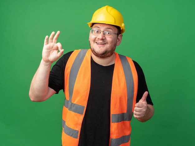 Homme de constructeur en gilet de construction et casque de sécurité regardant la caméra souriant joyeusement faisant signe ok montrant les pouces vers le haut debout sur fond vert