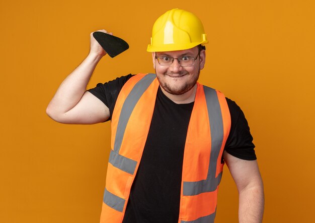 Homme de constructeur en gilet de construction et casque de sécurité tenant un couteau à mastic regardant la caméra en souriant avec un visage heureux