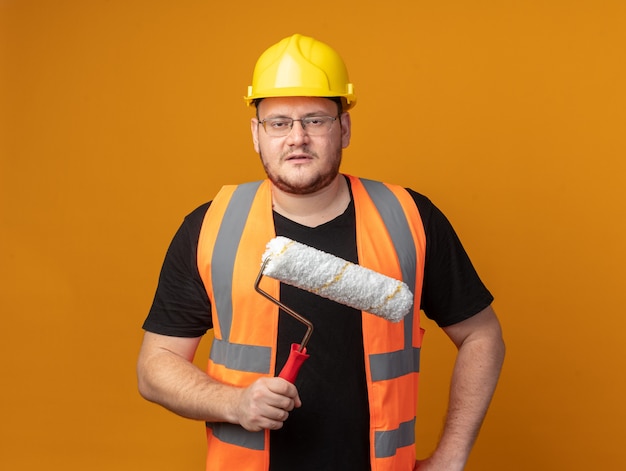 Homme de constructeur en gilet de construction et casque de sécurité tenant un rouleau à peinture regardant la caméra avec un visage sérieux