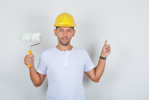 Homme constructeur montrant le mur et tenant le rouleau à peinture en t-shirt blanc, casque, vue de face