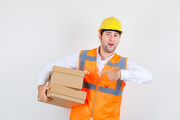 Homme de constructeur pointant le doigt sur des boîtes en carton en chemise, uniforme et à la recherche de positif. vue de face.