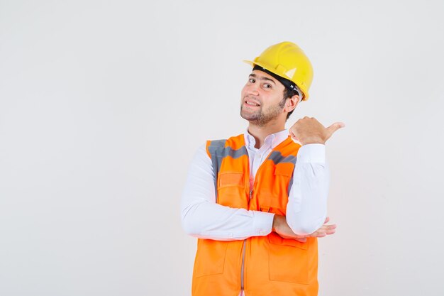 Homme de constructeur pointant le pouce vers le côté en chemise, uniforme et à la gaieté. vue de face.