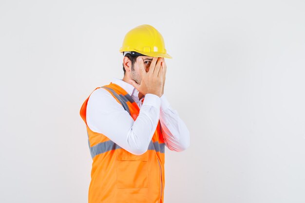 Homme de constructeur regardant à travers les doigts avec un œil en chemise, uniforme et à la gêne.