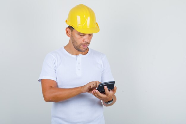 Homme de constructeur en t-shirt blanc, casque à l'aide de la calculatrice et regardant occupé, vue de face.