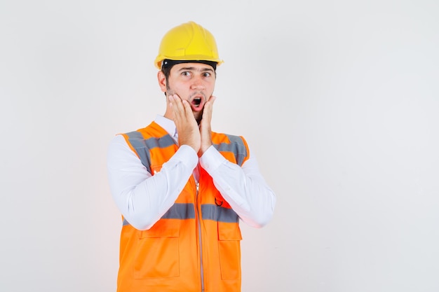 Homme de constructeur tenant la main près de la bouche en chemise, uniforme et à la recherche de choc. vue de face.