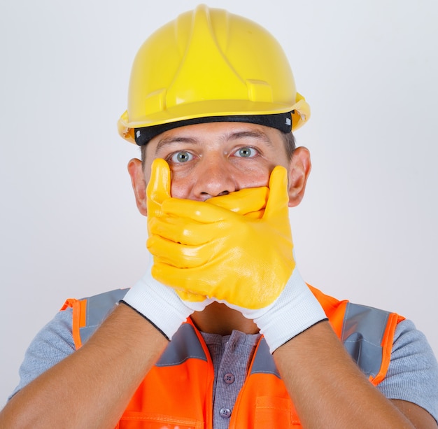 Homme constructeur en uniforme, casque, gants couvrant la bouche avec les mains pour erreur et à la vue choquée, de face.