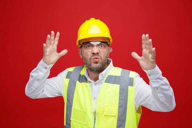 Homme constructeur en uniforme de construction et casque de sécurité portant des lunettes de sécurité en colère et frustré levant les bras se déchaîner debout sur fond rouge