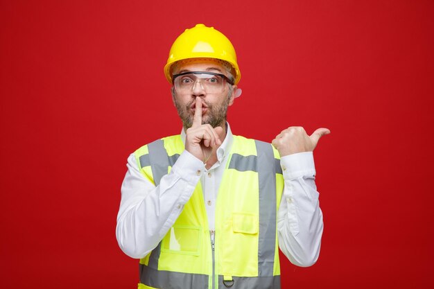 Homme constructeur en uniforme de construction et casque de sécurité portant des lunettes de sécurité regardant la caméra faisant un geste de silence avec le doigt sur les lèvres pointant avec le pouce sur le côté debout sur fond rouge