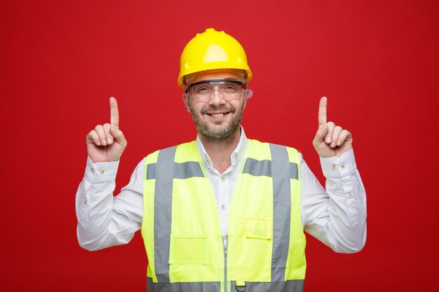 Homme constructeur en uniforme de construction et casque de sécurité portant des lunettes de sécurité regardant la caméra heureux et joyeux pointant avec l'index debout sur fond rose
