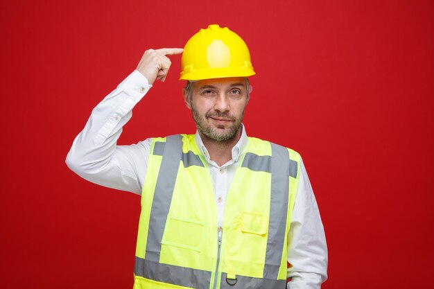 Homme constructeur en uniforme de construction et casque de sécurité regardant la caméra avec un sourire sceptique pointant avec l'index sur sa tête debout sur fond rouge