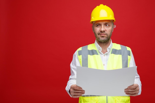 Homme constructeur en uniforme de construction et casque de sécurité tenant un plan regardant de côté avec une expression sceptique debout sur fond rouge