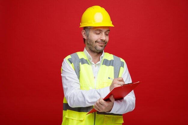 Homme constructeur en uniforme de construction et casque de sécurité tenant le presse-papiers à la confiance de prendre des notes debout sur fond rouge