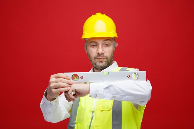 Homme constructeur en uniforme de construction et casque de sécurité tenant une règle de niveau à la confiance debout sur fond rouge