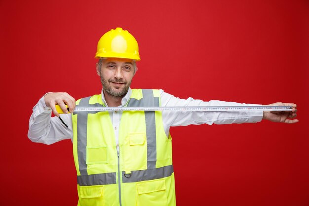 Homme constructeur en uniforme de construction et casque de sécurité tenant une règle regardant la caméra avec un sourire sur son visage debout sur fond rouge