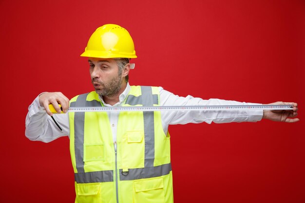 Homme constructeur en uniforme de construction et casque de sécurité tenant un ruban de mesure à la surprise debout sur fond rouge