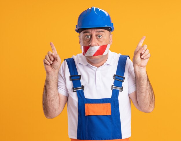 Homme de construction adulte caucasien choqué en uniforme couvre la bouche avec du ruban adhésif pointant vers le haut avec deux mains sur l'orange