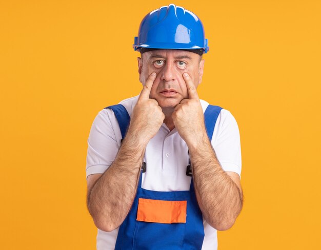 Homme de construction adulte caucasien confiant en uniforme tire les paupières sur orange