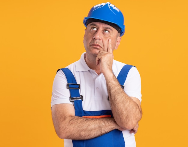 Homme de construction adulte caucasien réfléchi en uniforme met la main sur le menton et lève les yeux sur l'orange