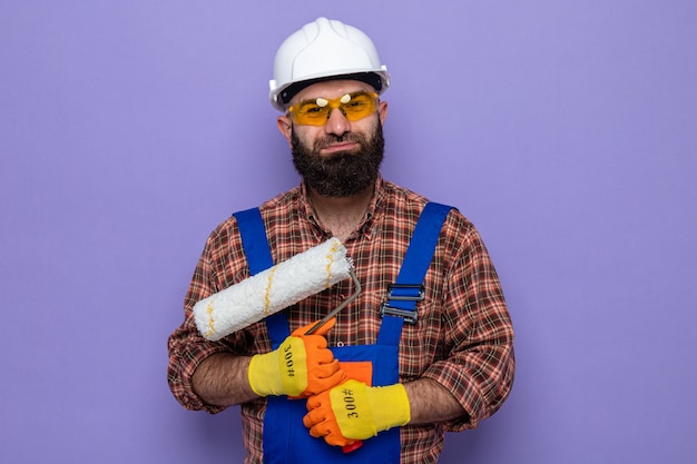 Homme de construction barbu en uniforme de construction et casque de sécurité portant des gants en caoutchouc tenant un rouleau à peinture à l'air heureux et souriant