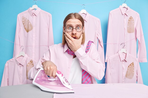 l'homme contre la bouche regarde effrayé a oublié une tâche de plus de la femme à propos des poses de la maison près de la planche à repasser caresse les vêtements bleus