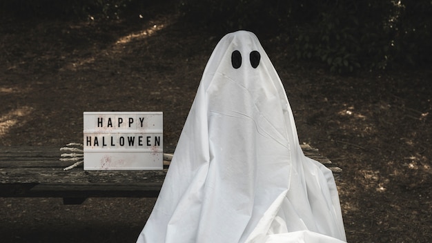 Photo gratuite homme en costume fantôme assis sur un banc près de la tablette d'halloween