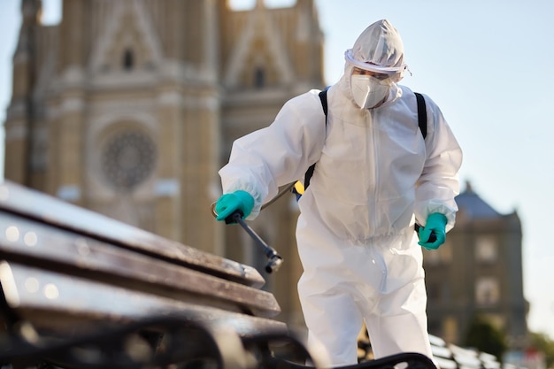 Photo gratuite homme en costume de matières dangereuses désinfectant un banc public dans la ville en raison de l'épidémie de coronavirus