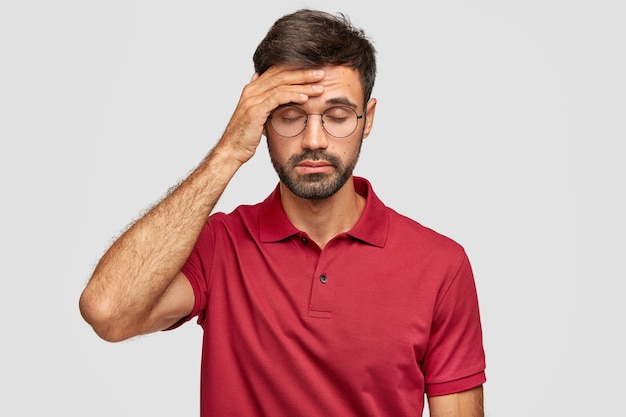 Un homme cuacasien surmené ressent de terribles maux de tête après une nuit sans sommeil, garde la main sur le front, ferme les yeux, vêtu d'un t-shirt rouge décontracté, se tient contre un mur blanc. Les gens et la fatigue