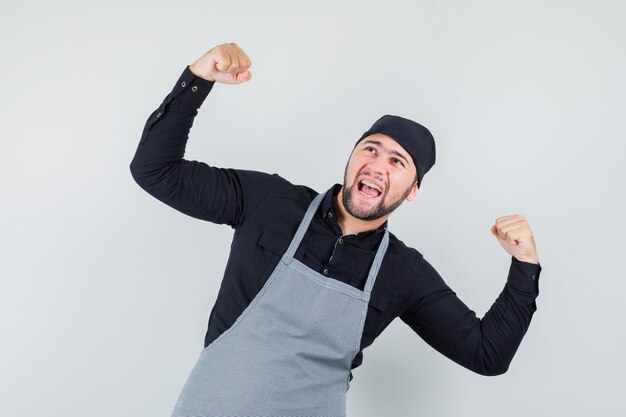 Homme cuisinier en chemise, tablier montrant le geste du gagnant et à la béatitude, vue de face.