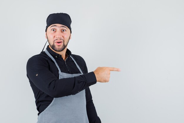 Homme cuisinier en chemise, tablier pointant vers le côté et à la vue étonnée, de face.