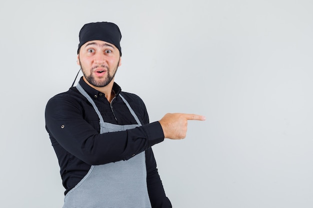 Homme cuisinier en chemise, tablier pointant vers le côté et à la vue étonnée, de face.