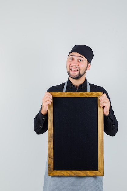 Homme cuisinier en chemise, tablier tenant tableau noir et à la joyeuse, vue de face.