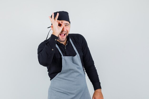 Homme cuisinier montrant signe ok sur les yeux en chemise, tablier et à la recherche amusée. vue de face.