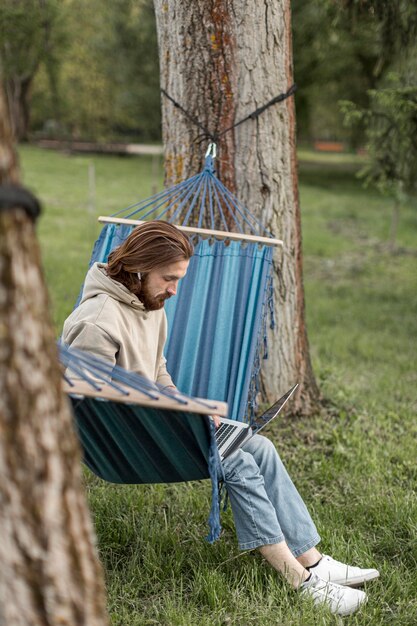 Homme dans la nature assis dans un hamac et travaillant sur ordinateur portable