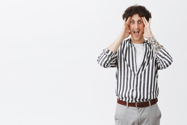 Homme dans une situation délicate en criant en essayant de ne pas regarder de côté tenant les paumes sur le visage en hurlant et en criant des yeux étonnés et étonnés debout dans une chemise et un pantalon rayés élégants sur un mur gris