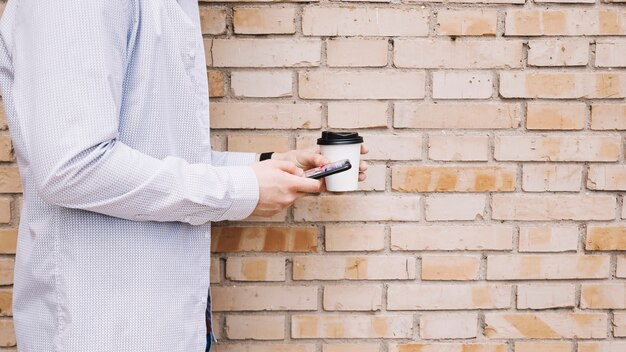 Homme, debout, devant, brique, mur, tasse café, et, téléphone portable, tenue, dans, main