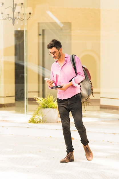 Photo gratuite homme debout à l'extérieur avec son sac à dos à l'aide d'un téléphone portable