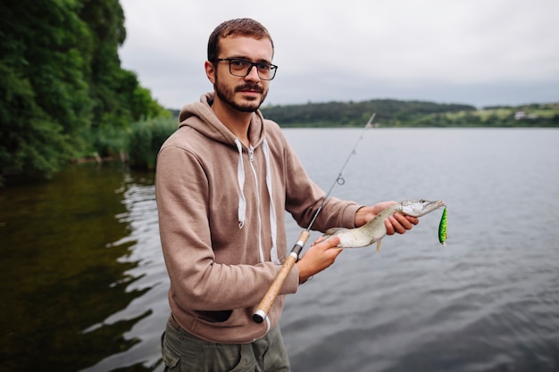 Photo gratuite homme debout près du lac tenant un poisson avec un crochet