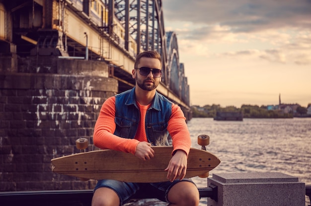 Photo gratuite homme décontracté avec longboard posant près de la rivière et du vieux pont ferroviaire.