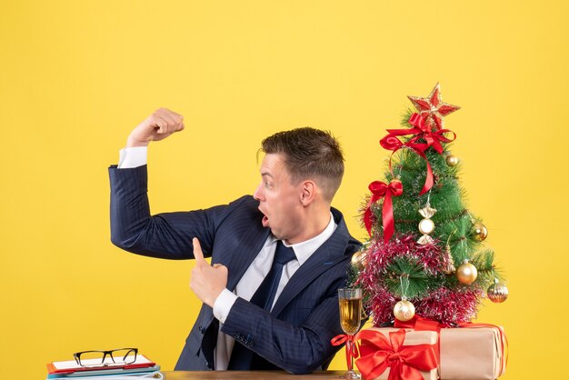 Homme demandé montrant son muscle assis à la table près de l'arbre de Noël et présente sur jaune