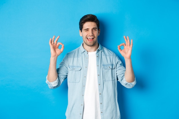 Homme détendu et confiant montrant des signes corrects et un clin d'œil, tout va bien, debout sur fond bleu.