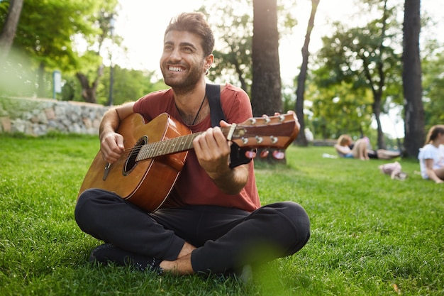 Homme détendu rêveur jouant de la guitare, s'asseoir sur l'herbe dans le parc avec instrument